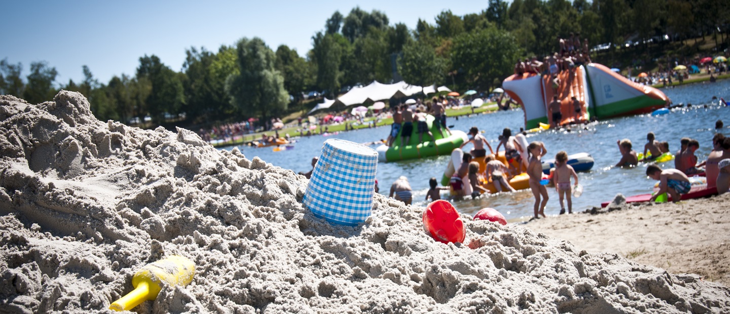 Kinderen en gezinnen genieten van een zonnige dag aan een officiële zwemlocatie in Hart van Limburg met zandstrand en speeltoestellen in het water.