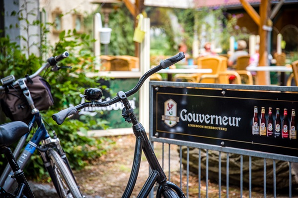 Twee fietsen geparkeerd bij een terras met een reclamebord van Brouwerij Lindeboom, met op de achtergrond mensen die genieten op het terras.
