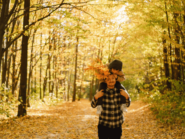 A man walks through the forest with a child sitting on his shoulders, holding leaves. The man and the child are looking at each other.