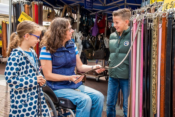 Drei Personen betrachten und diskutieren einen Gürtel an einem Marktstand auf dem Wochenmarkt in Weert. Eine Frau im Rollstuhl hält den Gürtel, während ein Junge und ein Mädchen zusehen und lachen. Im Hintergrund sind Marktstände mit Taschen und Accessoires zu sehen.