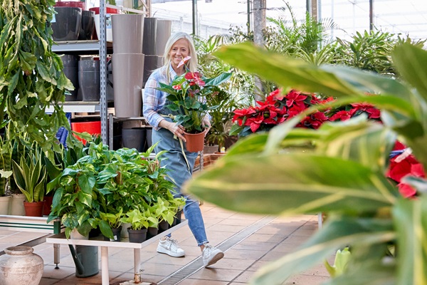 Frau in einem Gartencenter mit einem Blumentopf in der Hand, umgeben von grünen Pflanzen und Blumen.