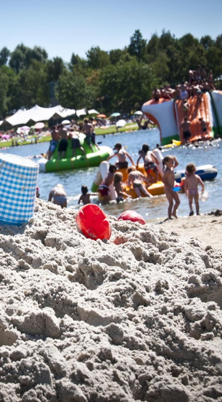 Kinderen en gezinnen genieten van een zonnige dag aan een officiële zwemlocatie in Hart van Limburg met zandstrand en speeltoestellen in het water.