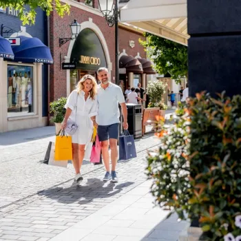 Stel wandelt met shoppingtassen door Designer Outlet Roermond op een zonnige dag, omringd door winkels en groen.