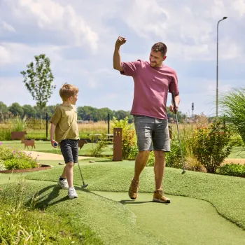 Vader viert een geslaagde slag met zijn zoon op de Adventure golfbaan van het park, omgeven door bloemen en gras.