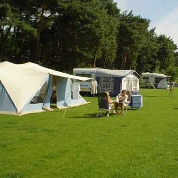 Kampeerders zitten bij hun tent en caravan op een grasveld van Camping Natuurplezier, met bos op de achtergrond.