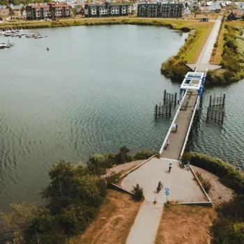 Brug over een Maasplas met wandelaars en fietsers, met woningen aan het water op de achtergrond.
