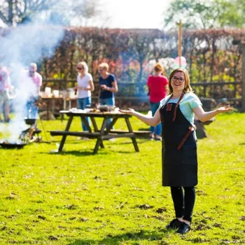 Vrouw in schort staat lachend op een grasveld tijdens een buitenkookworkshop, terwijl op de achtergrond wordt gekookt en gegeten.