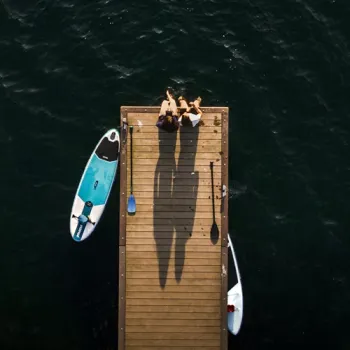 Twee mensen zitten op een houten steiger boven het water, met een supboard en peddels ernaast.