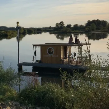 Vlot van Flossverleih TreibGuT ligt aangemeerd op de Maasplassen, terwijl twee mensen op het dakterras uitkijken over het water.