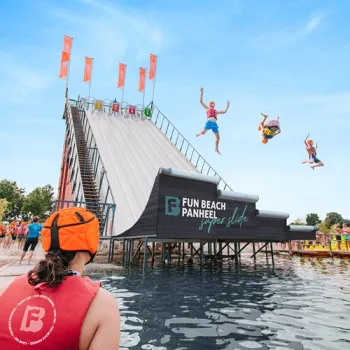 Mensen springen vanaf een hoge glijbaan bij Fun Beach Panheel het water in, terwijl toeschouwers vanaf de kant meekijken.