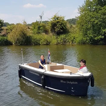 Twee mensen varen in een sloep over de Maas, omringd door groen langs de oever.