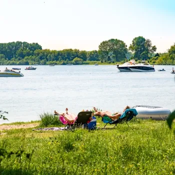 Mensen genieten van de zon aan de waterkant, terwijl boten en zeiljachten over de Maasplassen varen.