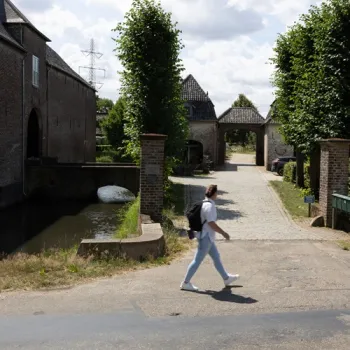 Persoon loopt langs de ingang van een hoeve met water, bomen en een poort op de achtergrond.
