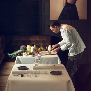 Chef presenteert een gerecht in Restaurant Damianz in Roermond, in een stijlvolle setting met gedekte tafels en ingrediënten op de voorgrond.