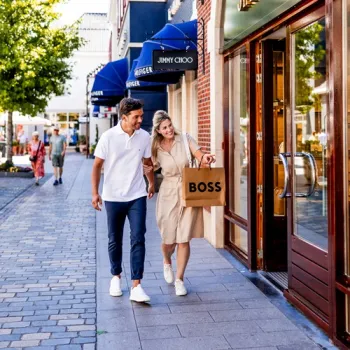 Stel loopt langs winkels in Designer Outlet Roermond met een Boss-tas, terwijl ze winkelen in een levendige straat met bekende merken.