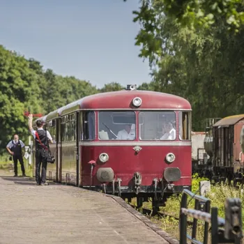 Historische rode trein op een perron met conducteur en passagiers, klaar voor vertrek in een groene omgeving.
