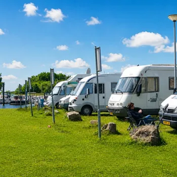 Camperplaats Hanssum aan de Maas in Neer met een rij campers op een zonnige zomerdag. Op de voorgrond ontspant een campergast in een stoel, terwijl op de achtergrond de jachthaven en boten zichtbaar zijn. De groene grasstrook en de blauwe lucht zorgen voor een rustige sfeer aan het water.